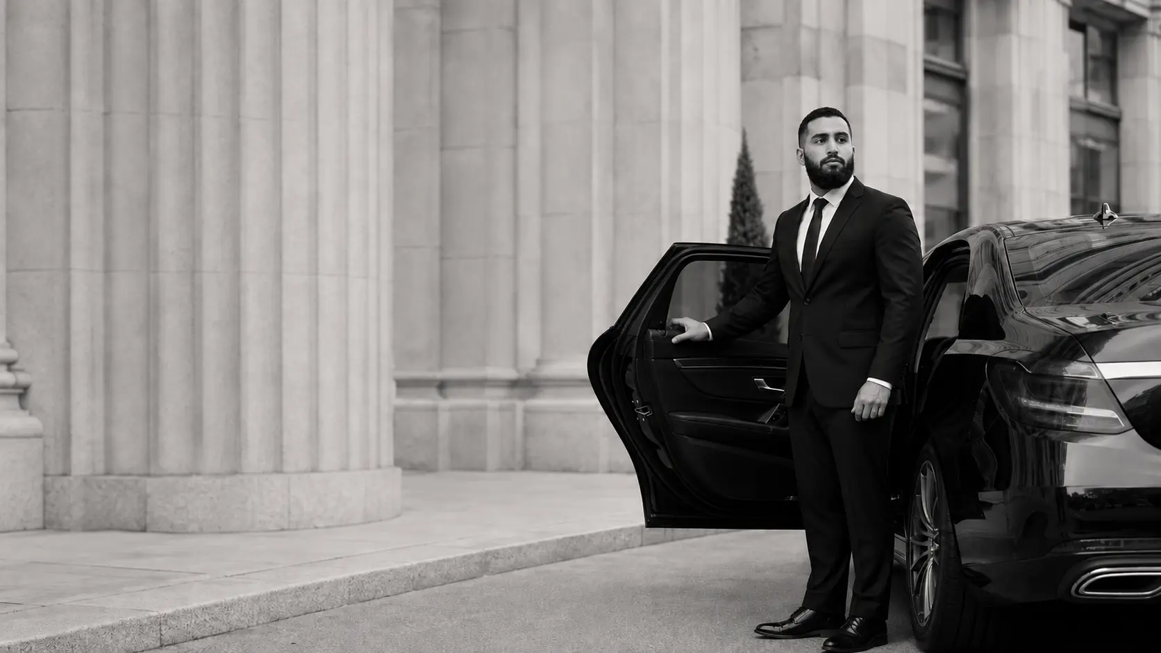 Fahad Hamid in a tailored black suit holding open the rear door of a black sedan